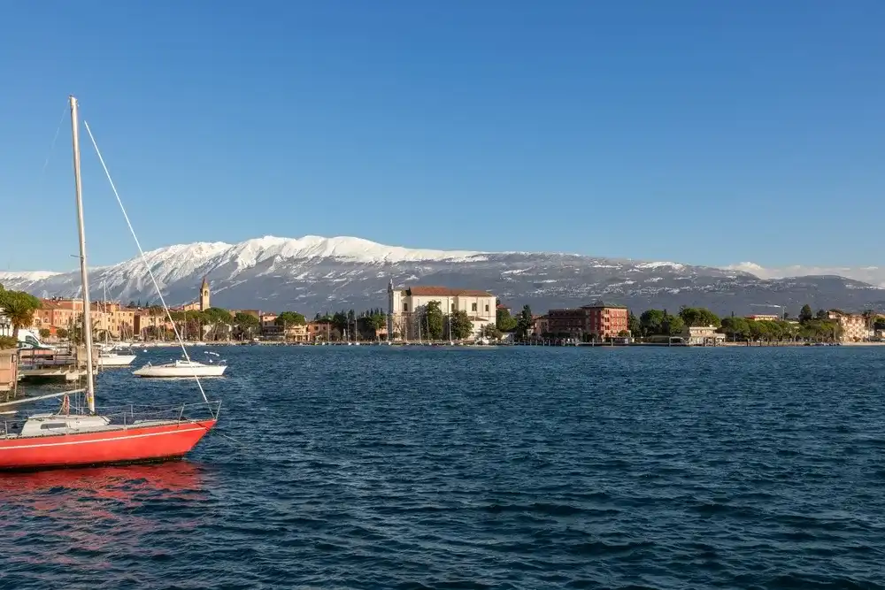 Embankment of the town Moniga del Garda on lake Garda against the background of the Alpine mountains with snow-capped peaks Embankment of the town Moniga del Garda on lake Garda against the background of the Alpine mountains with snow-capped peaks