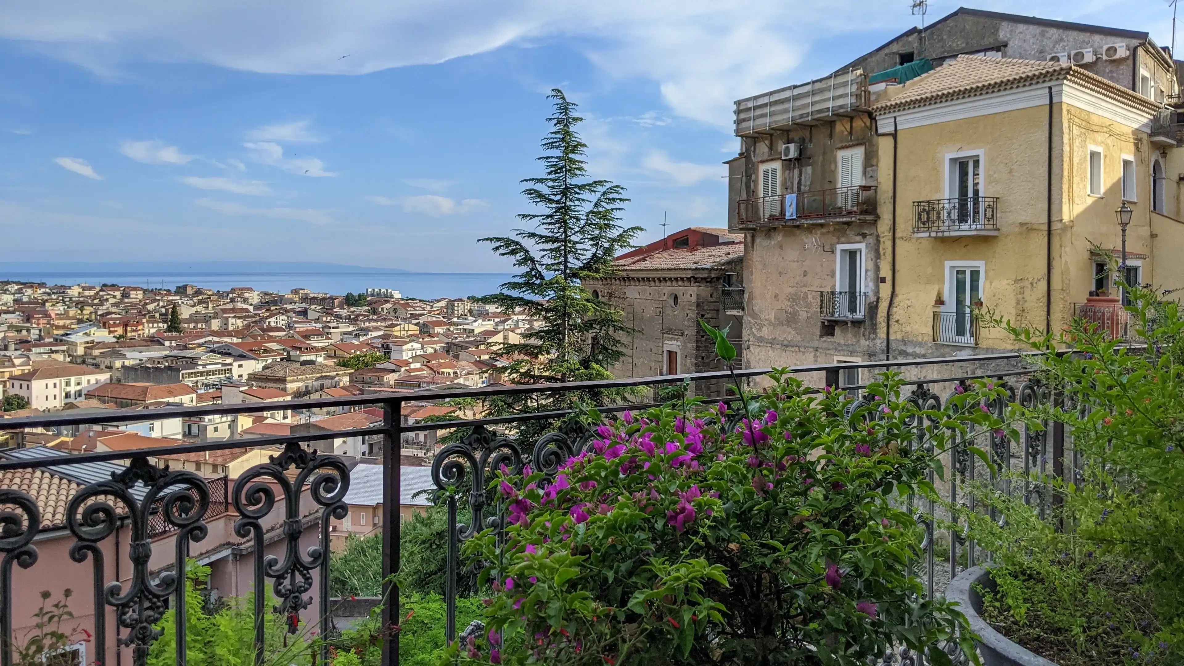 Amantea, Cosenza, Italy - July 4, 2021:Scenic overlook of Mediterranean town with terracotta roofs, ornamental iron balcony railing, flowering plants and cypress tree Amantea, Cosenza, Italy - July 4, 2021:Scenic overlook of Mediterranean town with terracotta roofs, ornamental iron balcony railing, flowering plants and cypress tree