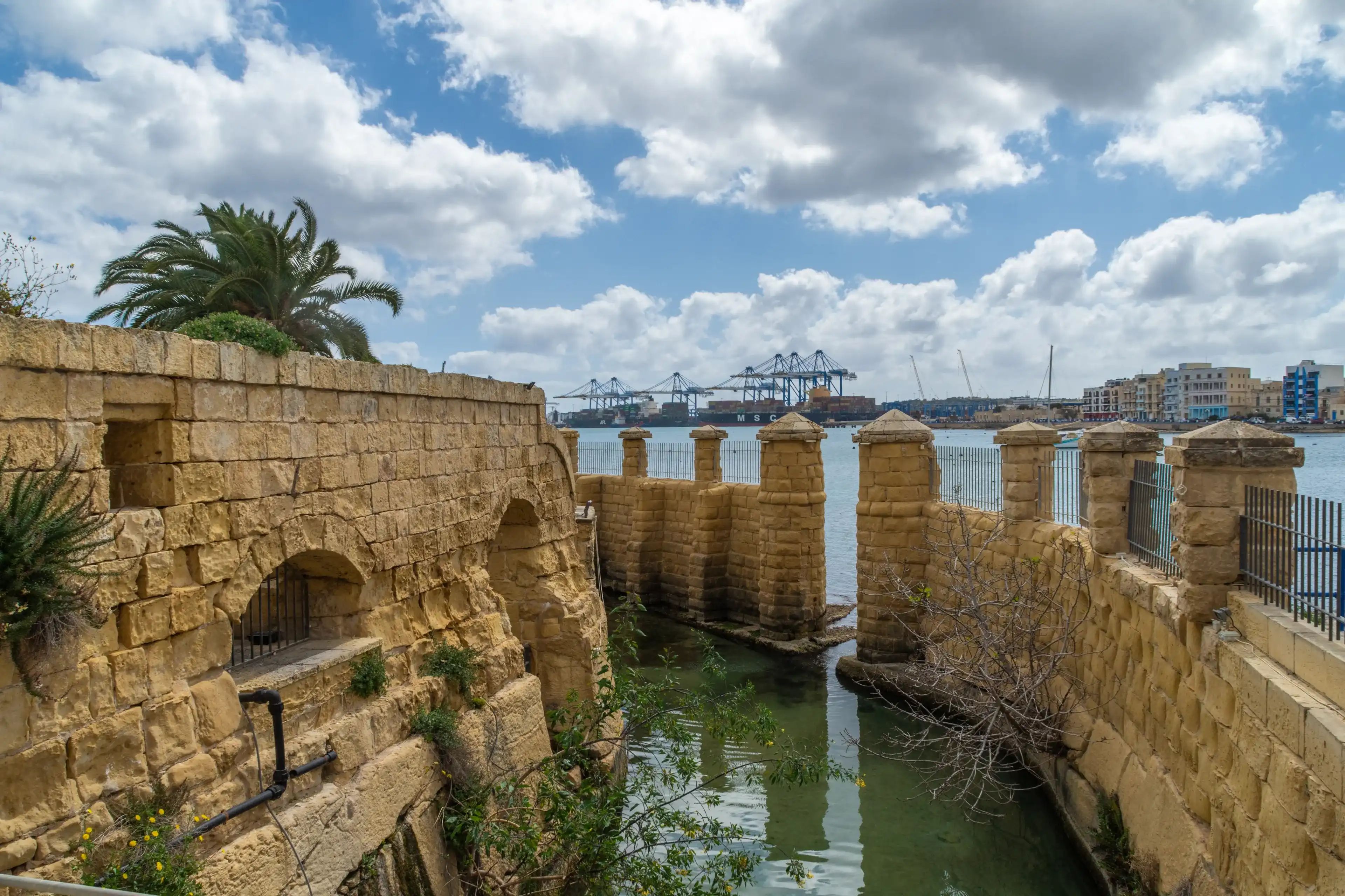 Birżebbuġa, Malta - March 14th 2021: The moat around the Ferretti Battery built by the Order of Saint John in 1716. The Malta Freeport can be seen in the background. Birżebbuġa, Malta - March 14th 2021: The moat around the Ferretti Battery built by the Order of Saint John in 1716. The Malta Freeport can be seen in the background.