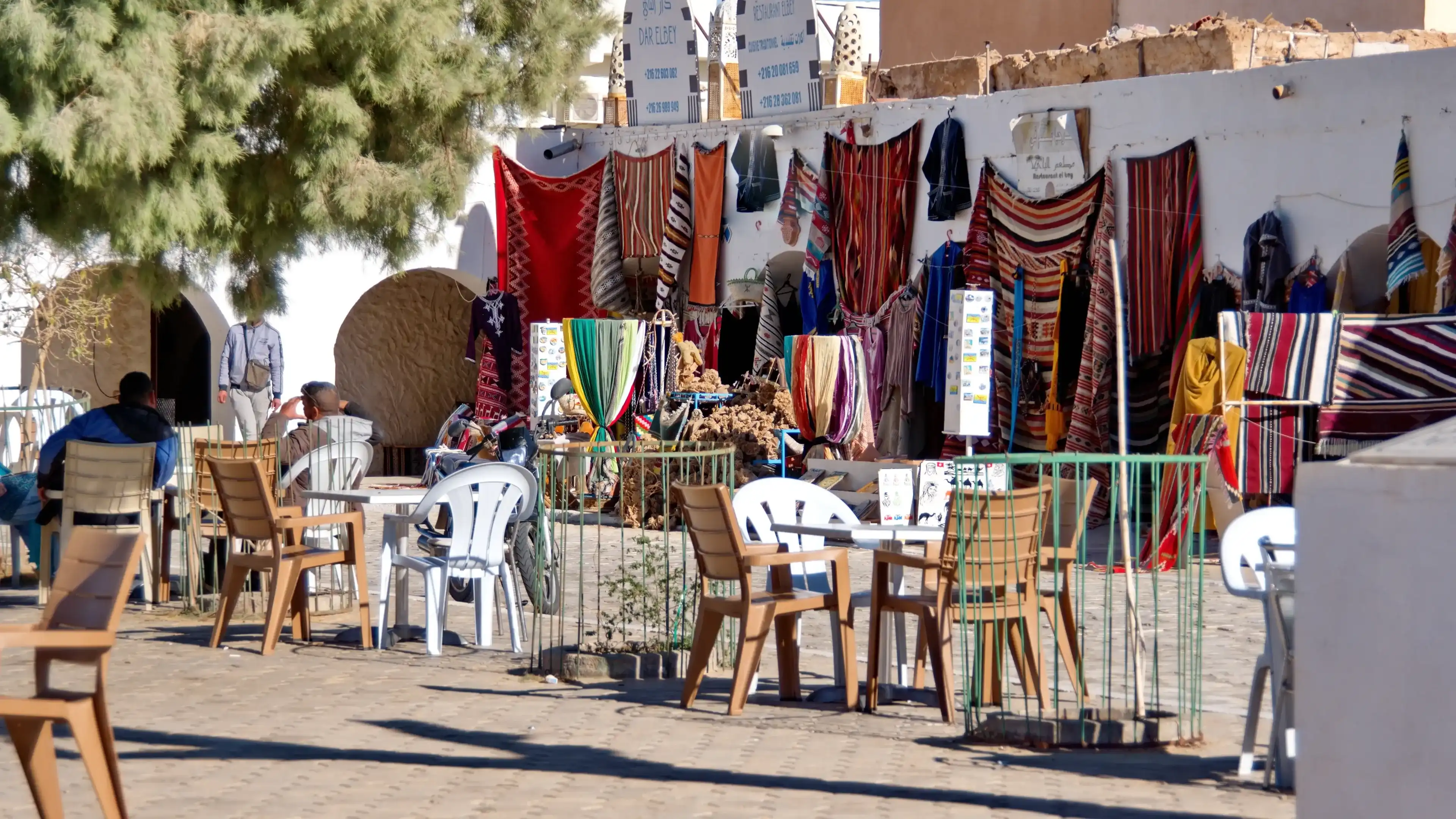 Douz, Tunisia - March 5, 2023: Cafe, with tables and chairs in the courtyard, set up in front of a stall selling colorful textiles in the market Douz, Tunisia - March 5, 2023: Cafe, with tables and chairs in the courtyard, set up in front of a stall selling colorful textiles in the market