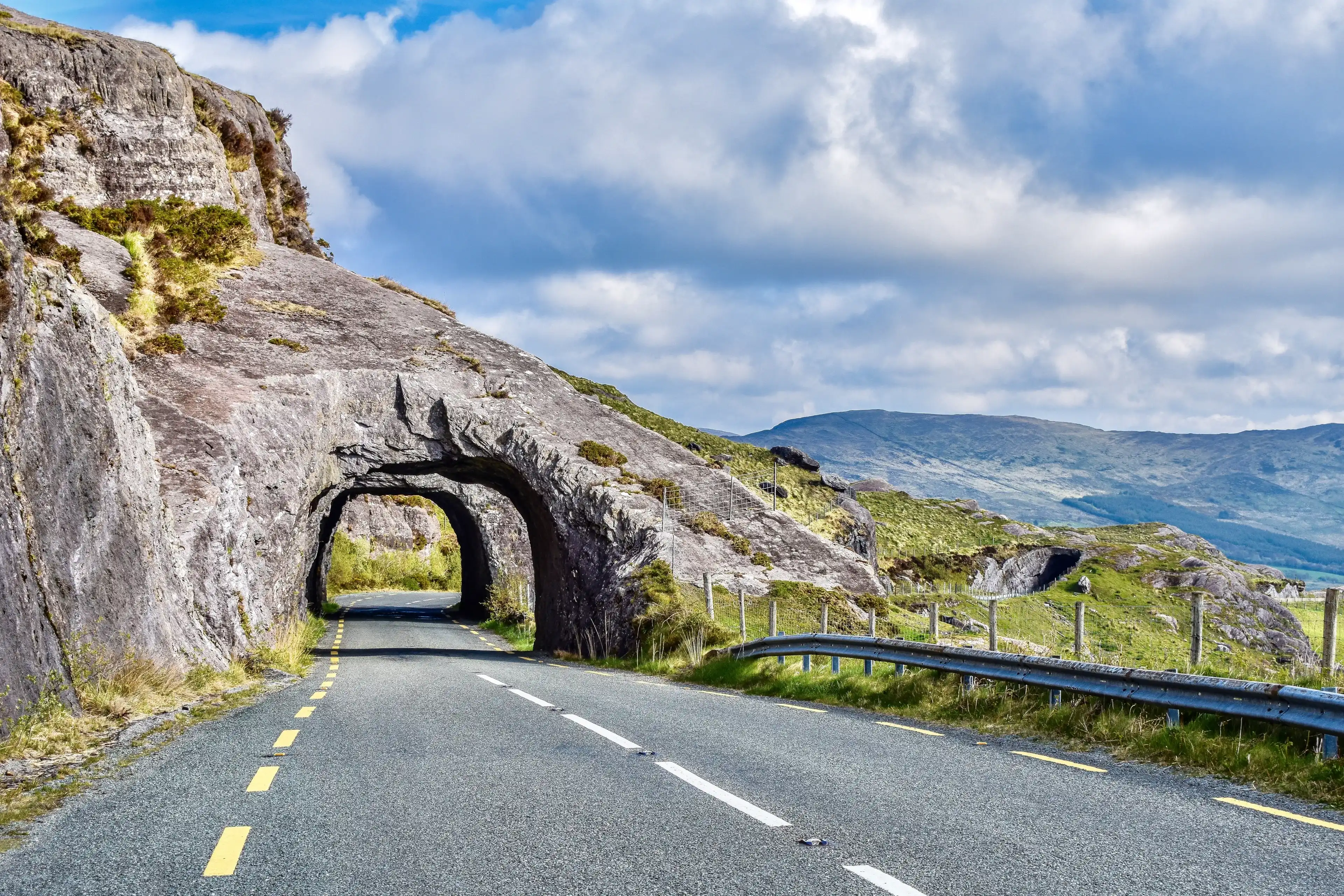Kenmare Road, County Kerry, Ireland, stone arch tunnels made from gray stone showing lined highway N71 with gray guardrail in the foreground backed by light green mountains and a mostly cloudy sky. Kenmare Road, County Kerry, Ireland, stone arch tunnels made from gray stone showing lined highway N71 with gray guardrail in the foreground backed by light green mountains and a mostly cloudy sky.