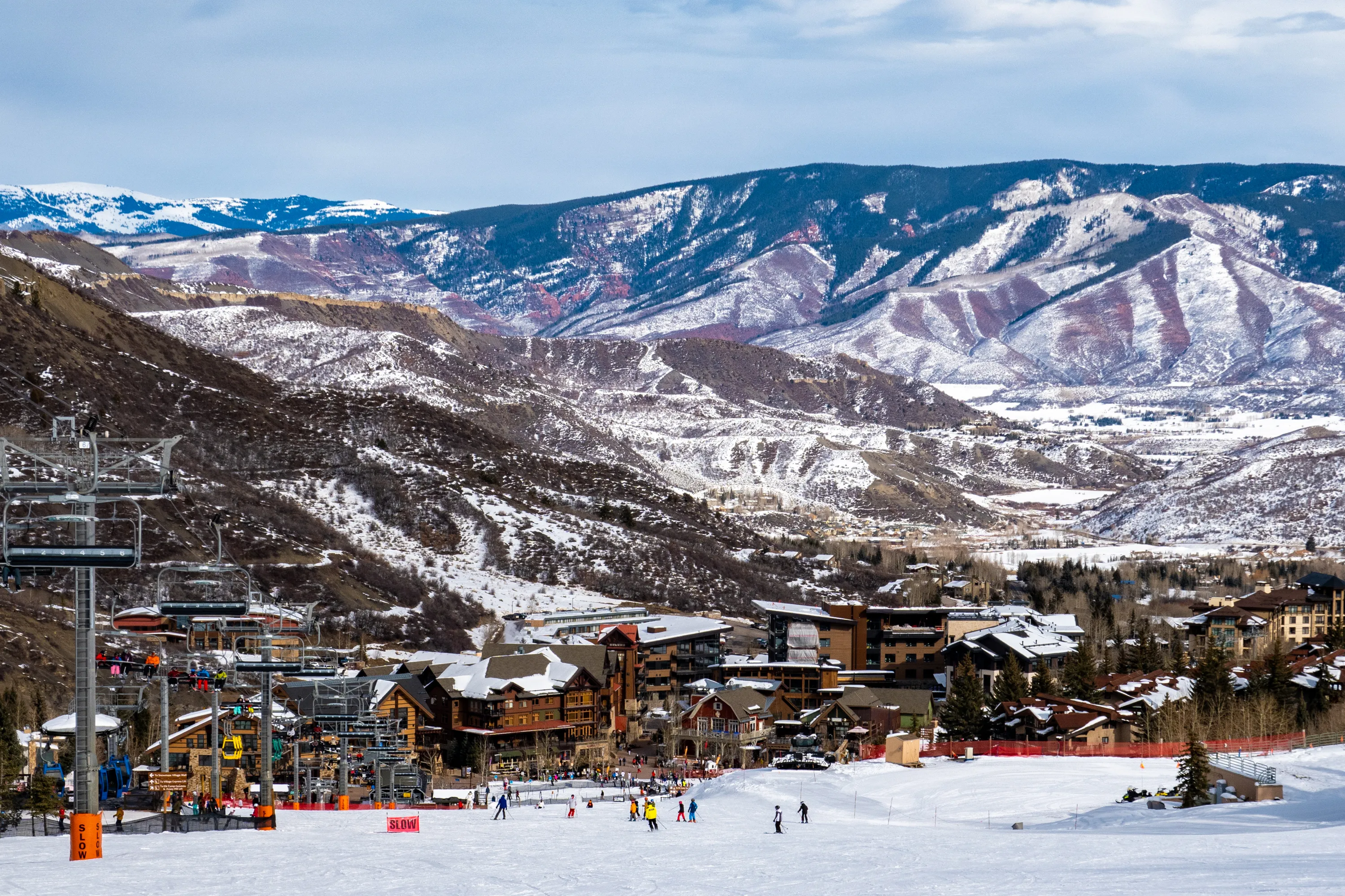 Panoramic view of Snowmass Village, with skiers skiing at the Aspen Snowmass ski resort in the foreground and the Rocky Mountains of Colorado in the background, on a partly cloudy winter day. 