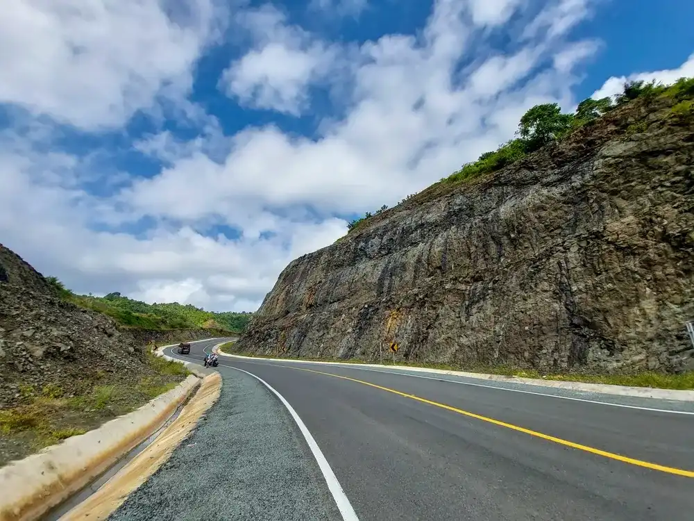 Scenic view of the Southern Cross Route in Tulungagung, Indonesia. A smooth winding asphalt road cutting through rocky hills under a bright blue sky with fluffy clouds. Scenic view of the Southern Cross Route in Tulungagung, Indonesia. A smooth winding asphalt road cutting through rocky hills under a bright blue sky with fluffy clouds.