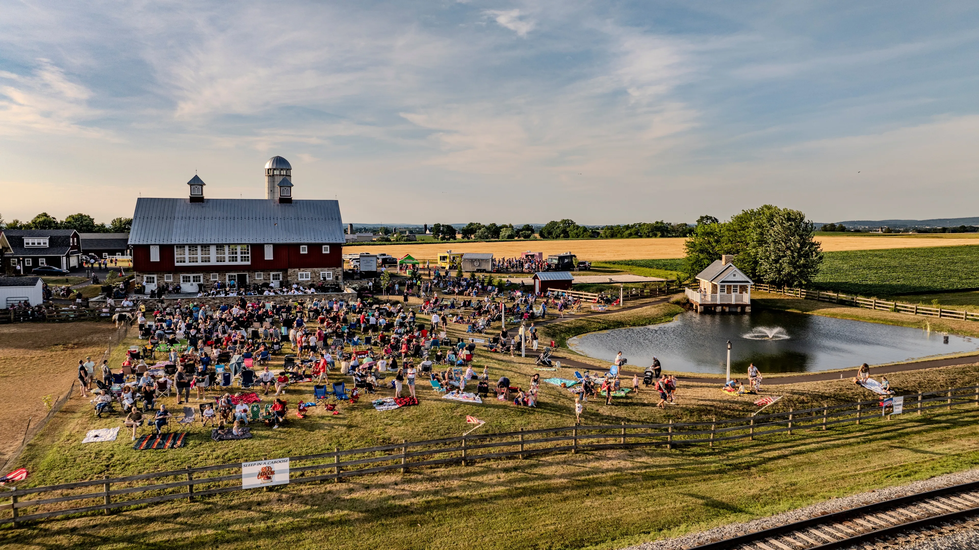 Ronks, Pennsylvania, USA, July 3, 2024 - Large Crowd Gathered On A Farm Near A Red Barn And A Small Pond With Fountains, Enjoying An Outdoor Event On A Sunny Day.