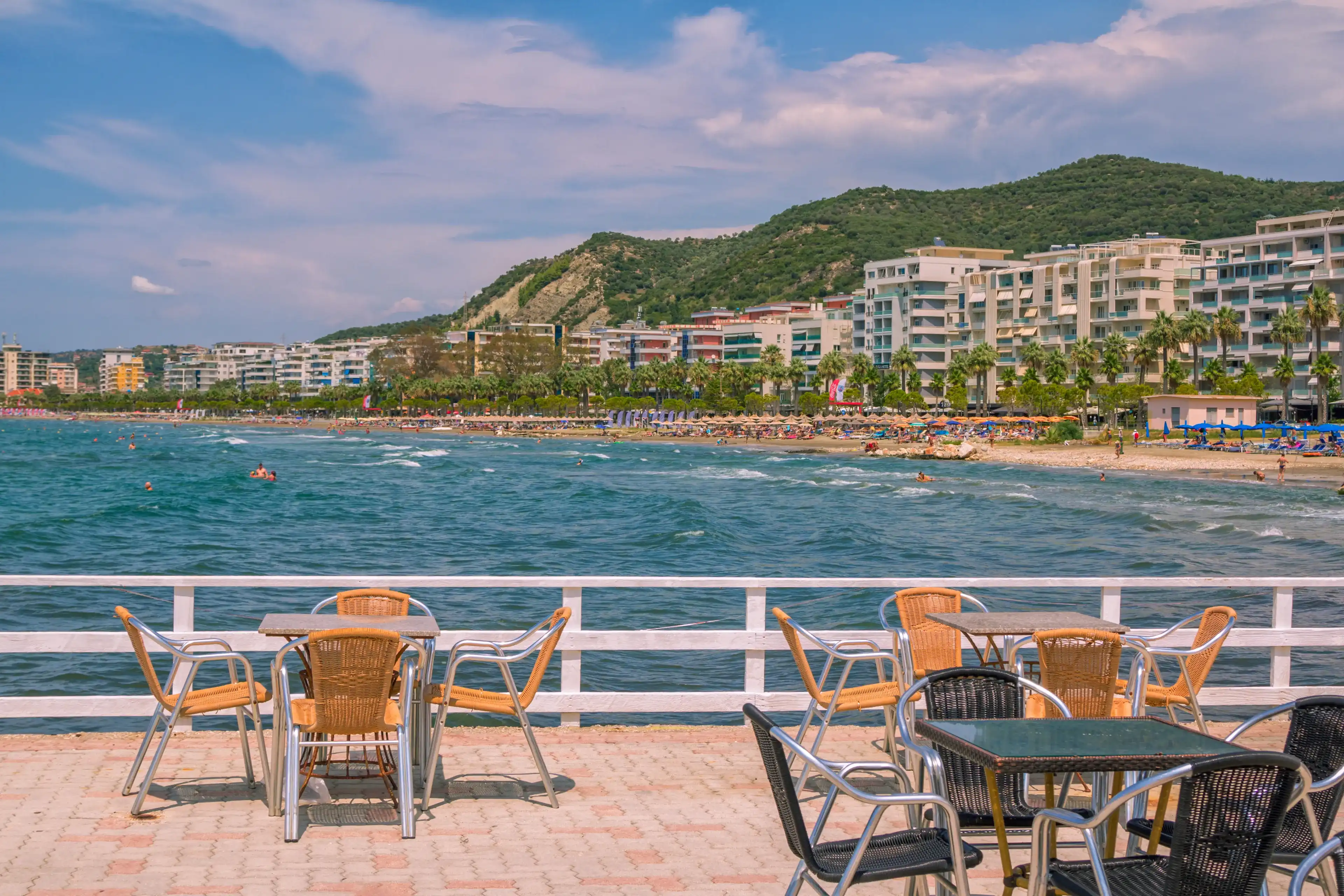 Vlora, Albania - August 7, 2020: summer resort –outdoors café – tables and chairs, turquoise sea water, people swimming, sunbathing on the beach, hotels buildings, blue sky and mountain on background Vlora, Albania - August 7, 2020: summer resort –outdoors café – tables and chairs, turquoise sea water, people swimming, sunbathing on the beach, hotels buildings, blue sky and mountain on background