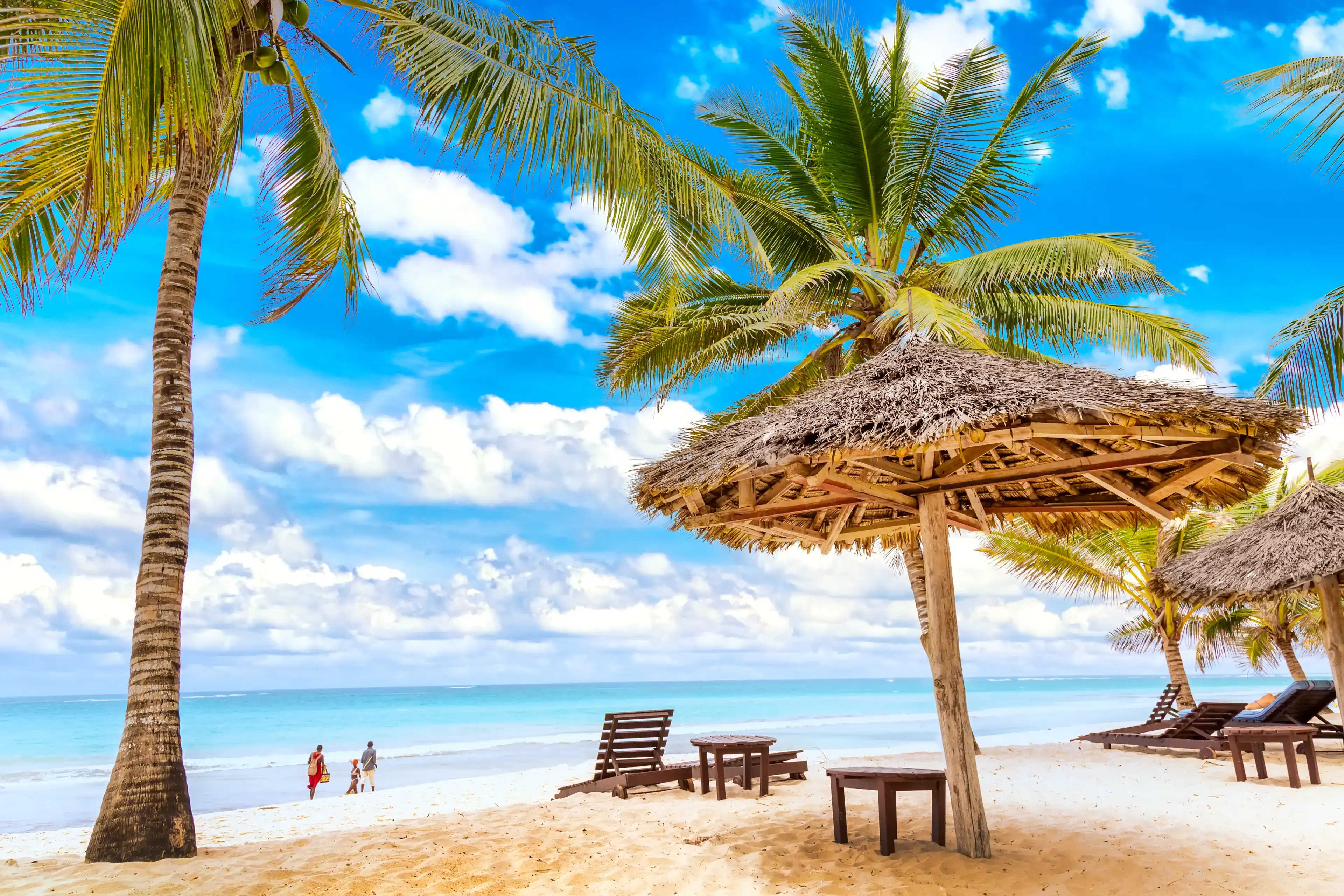 Sun loungers under umbrella and palms on the sandy beach by the ocean and cloudy sky. Vacation background. Idyllic beach landscape in Diani beach, Kenya, Africa. Sun loungers under umbrella and palms on the sandy beach by the ocean and cloudy sky. Vacation background. Idyllic beach landscape in Diani beach, Kenya, Africa.