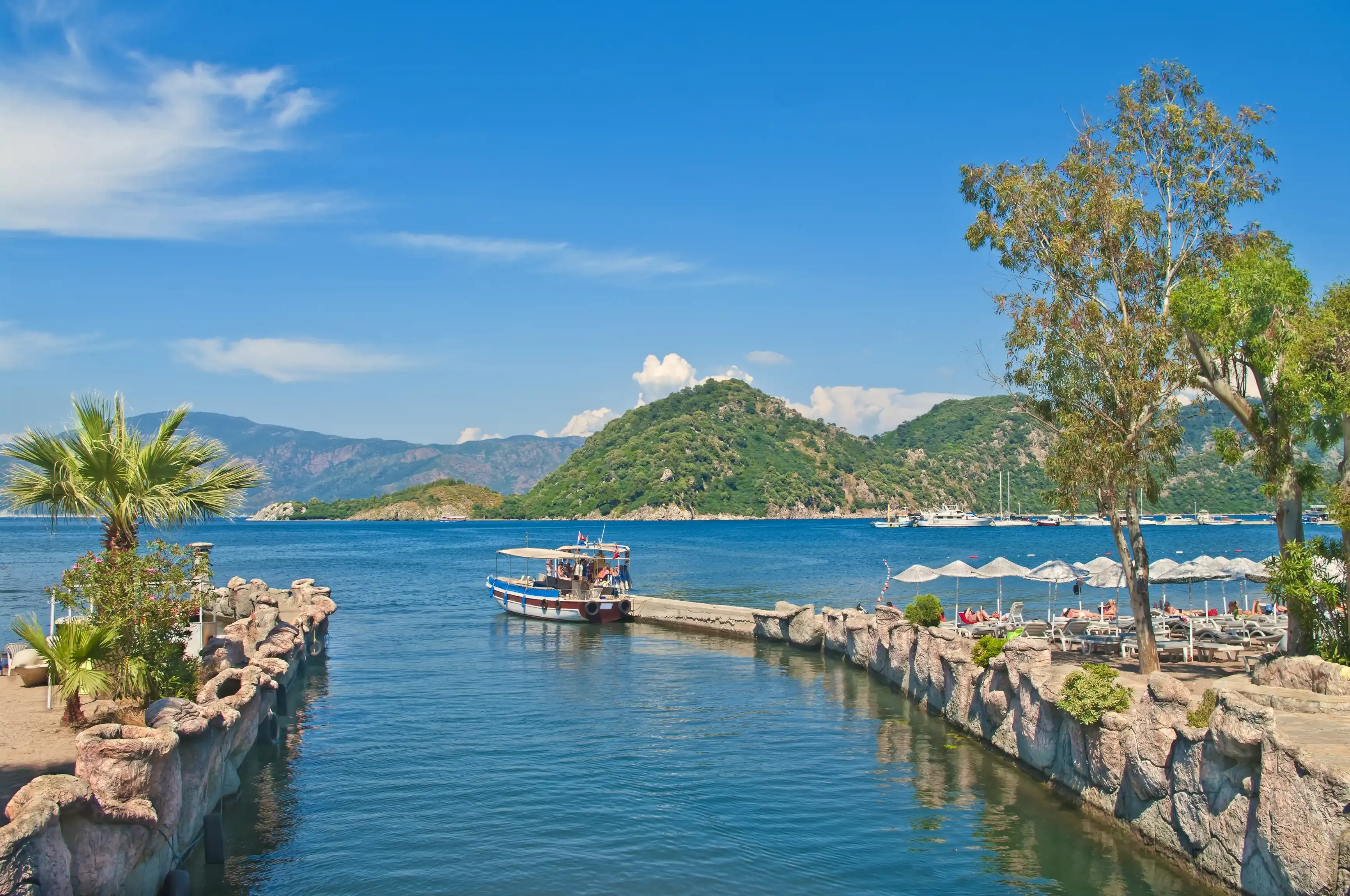 small tourist boat at Icmeler pier in Aegean sea bay with palm trees at foreground and mountains at background, Marmaris, Turkey small tourist boat at Icmeler pier in Aegean sea bay with palm trees at foreground and mountains at background, Marmaris, Turkey