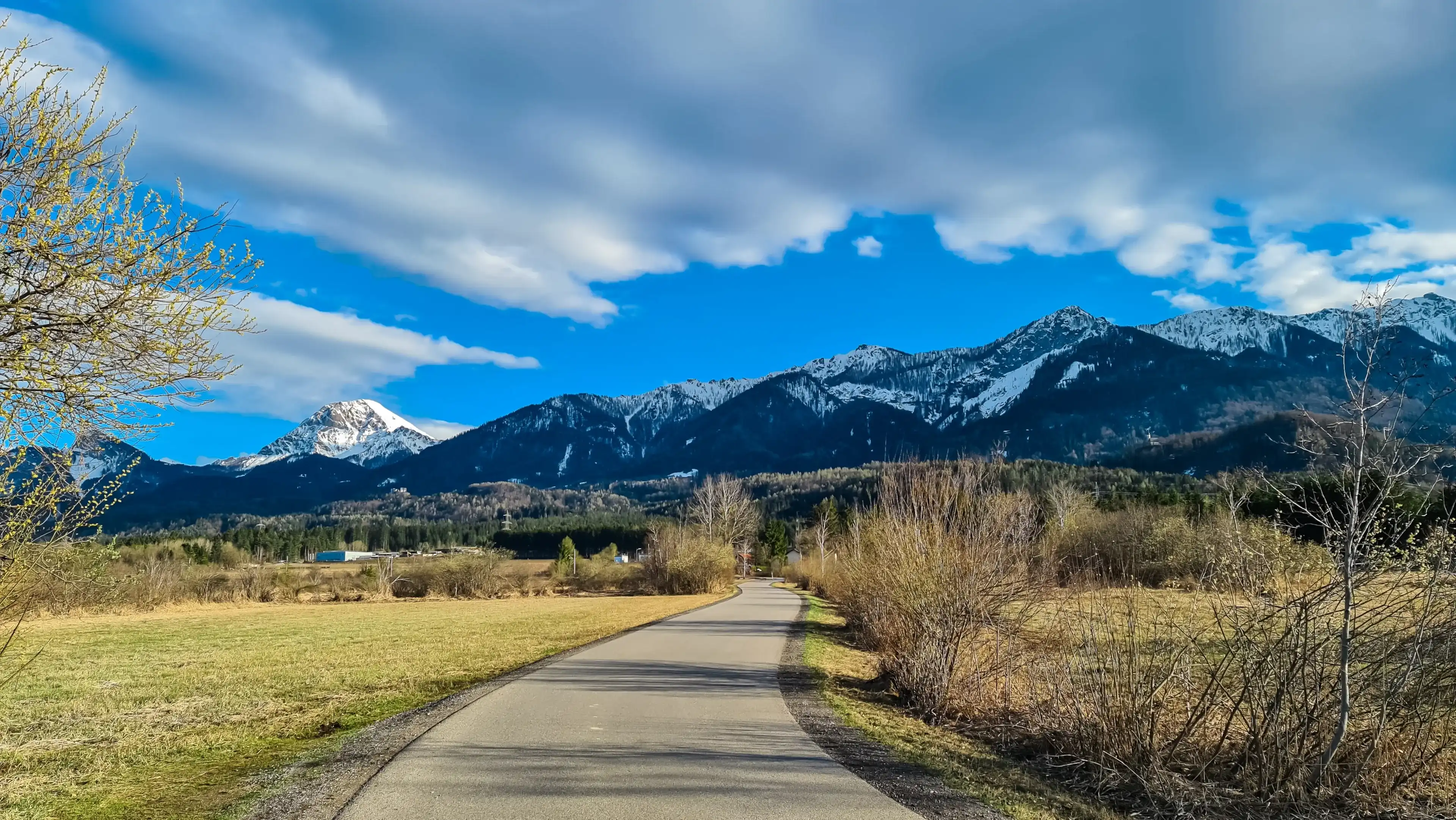 Paved road with scenic view of majestic snow capped mountain peak Mittagskogel, Karawanks, Carinthia, Austria. Landscape of Finkensteiner Moor covered by grass and reed. Idyllic hiking trail in spring Paved road with scenic view of majestic snow capped mountain peak Mittagskogel, Karawanks, Carinthia, Austria. Landscape of Finkensteiner Moor covered by grass and reed. Idyllic hiking trail in spring