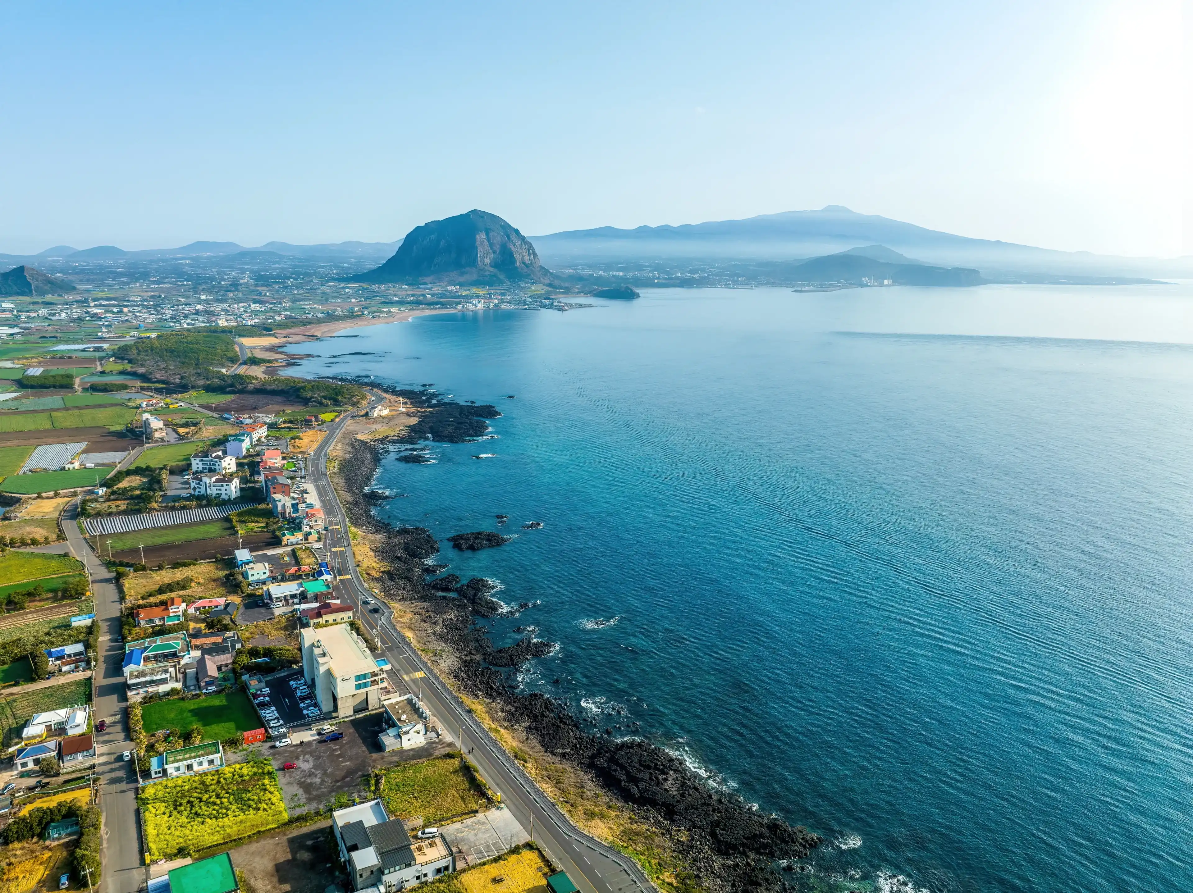 Daejeong-eup, Seogwipo-si, Jeju-do, South Korea - March 16, 2022: Aerial and morning view of road and houses of a village near Sagye Beach against Sanbangsan and Hallasan Mountain in the backgroud Daejeong-eup, Seogwipo-si, Jeju-do, South Korea - March 16, 2022: Aerial and morning view of road and houses of a village near Sagye Beach against Sanbangsan and Hallasan Mountain in the backgroud